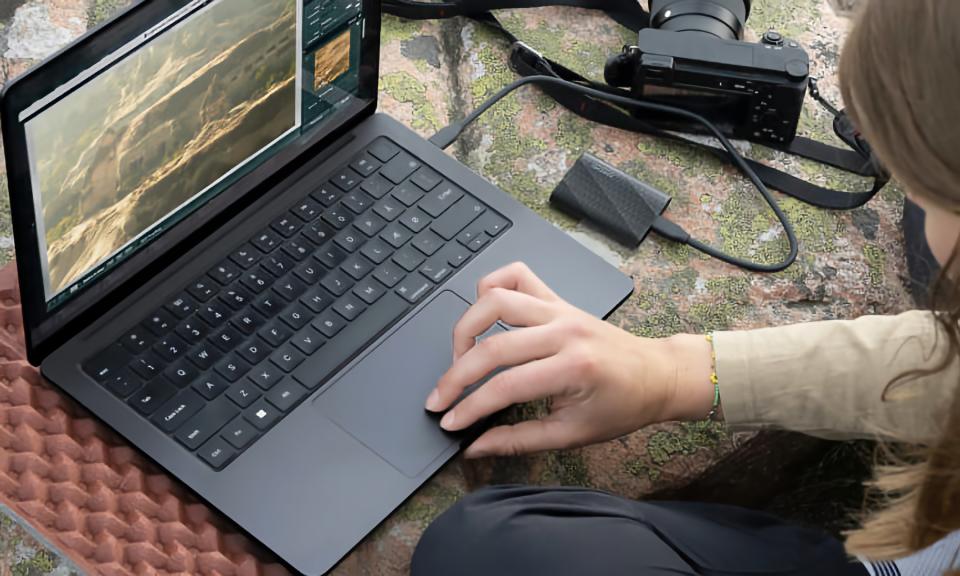 Lifestyle marketing photo of a person using a Windows laptop with a Samsung portable SSD plugged into it. A camera is also nearby, and it all sits on a green and orange surface.