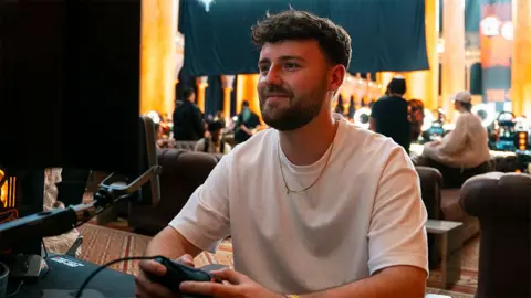 OllMS OllMS, a male CoD content creator, wearing a white tshirt while playing on a games console holding a black controller and looking into a screen while smiling. Behind him are several other gamers sitting on sofas.