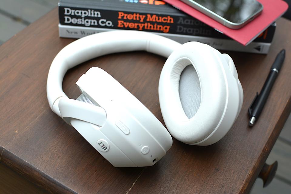 White headphones sitting on a wooden table from above with a red notebook and phone in the background sitting on top of a black and a white book.