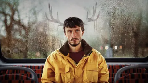 Netflix Promotional image for Netflix show Baby Reindeer, featuring actor and comedian Richard Gadd sitting on the back seat of a bus with antlers drawn in condensation on the window behind him.