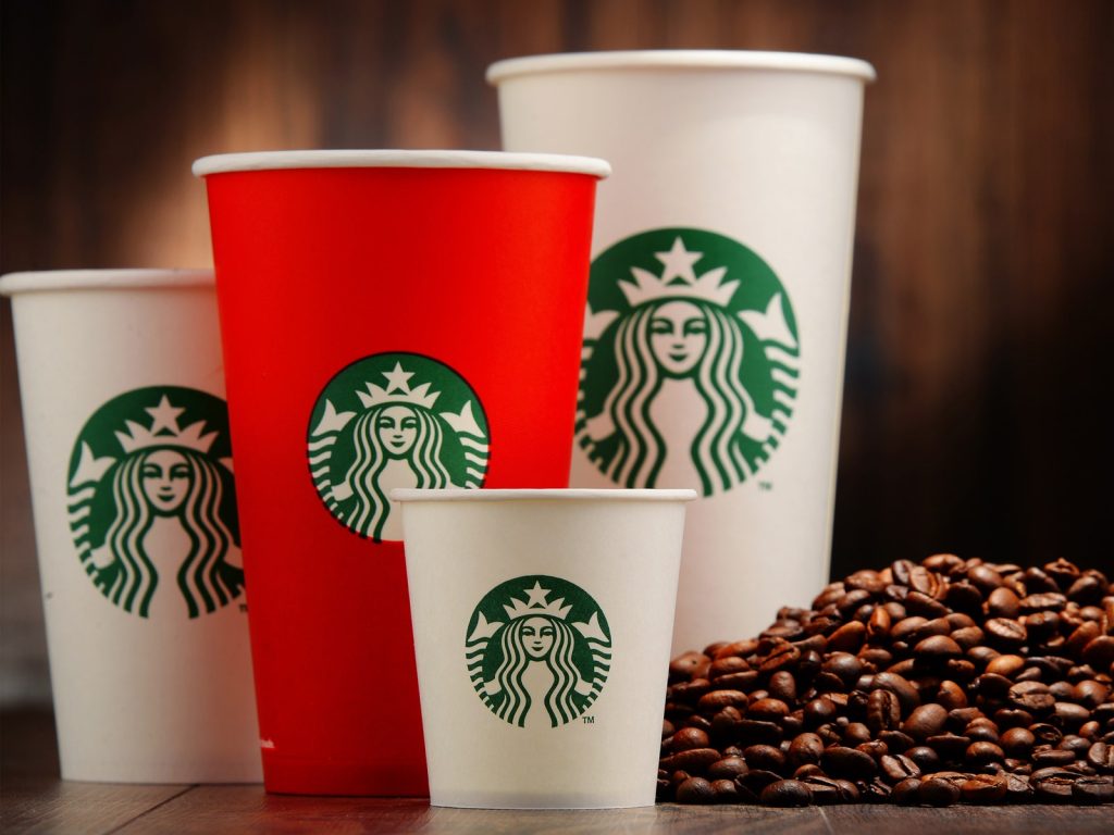 3 cốc giấy Starbucks trắng và 1 cốc giấy màu đỏ trên bàn màu nâu bên cạnh một đống hạt cà phê