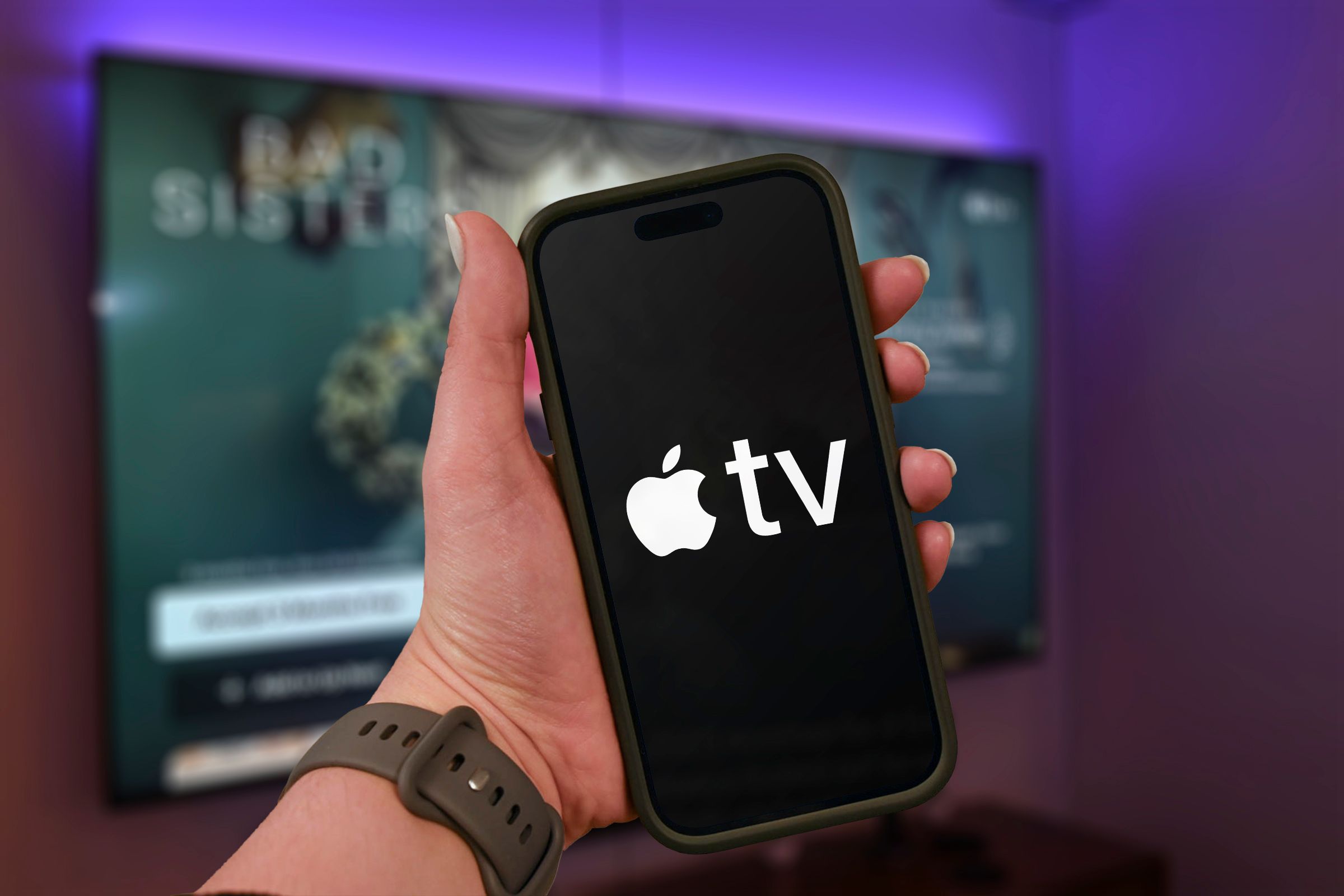 Tay cầm điện thoại có logo Apple TV+ và TV mờ ở chế độ nền.